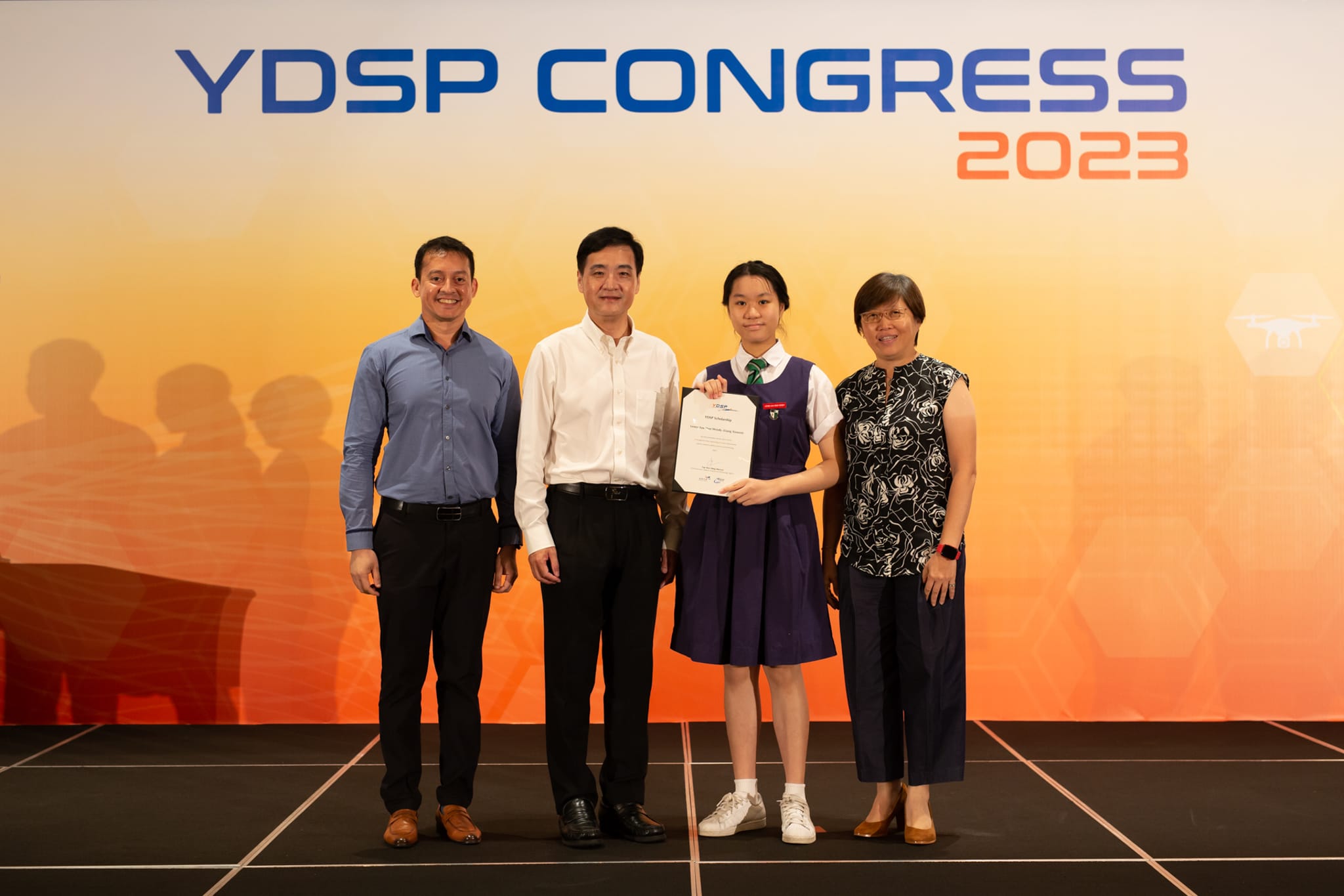Four people stand before a YDSP Congress 2023 banner; a young girl in school uniform holds a certificate.